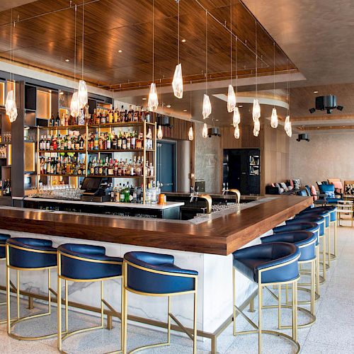 A modern bar with blue stools, a well-stocked liquor display, pendant lights, and a cozy seating area in a stylish lounge.