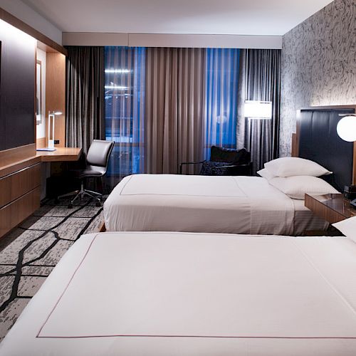 A modern hotel room with two beds, a wall-mounted TV, a desk by the window, and stylish lighting, creating a cozy atmosphere.