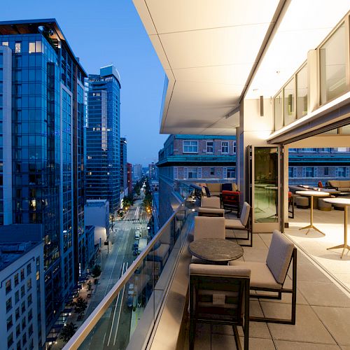A modern rooftop terrace with tables and chairs overlooks a city street lined with tall buildings, captured at dusk.