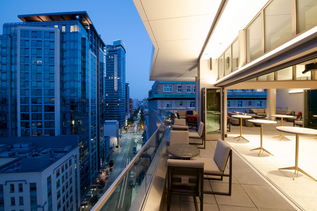 City view from a rooftop terrace at dusk, featuring modern buildings, outdoor seating, and warm lighting.