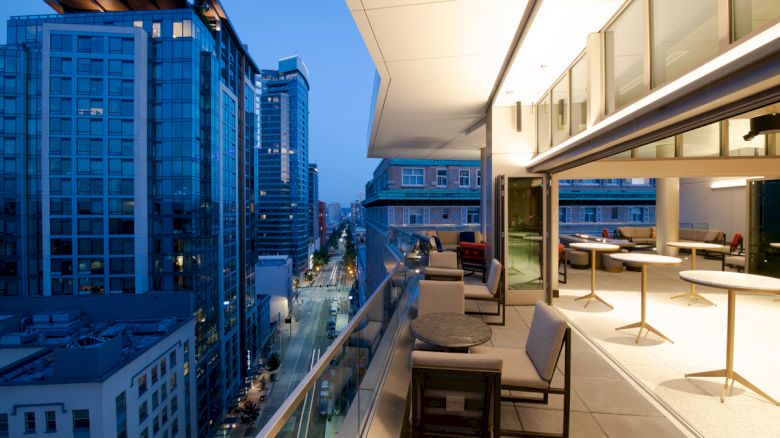 City view from a rooftop terrace at dusk, featuring modern buildings, outdoor seating, and warm lighting.