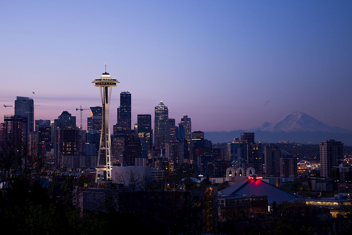 The image shows a skyline with the Space Needle in Seattle at dusk, along with city buildings and Mount Rainier in the background.