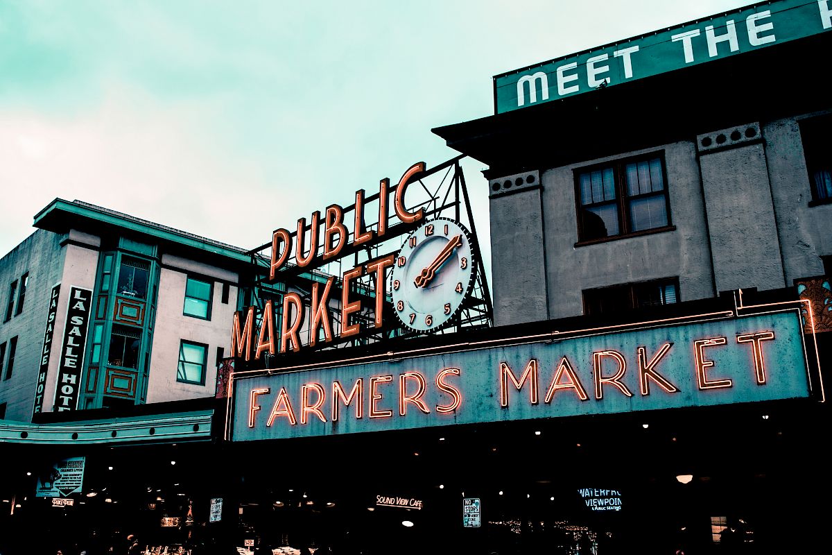 The image shows a public market sign with a clock and a neon "Farmers Market" sign in an urban setting.