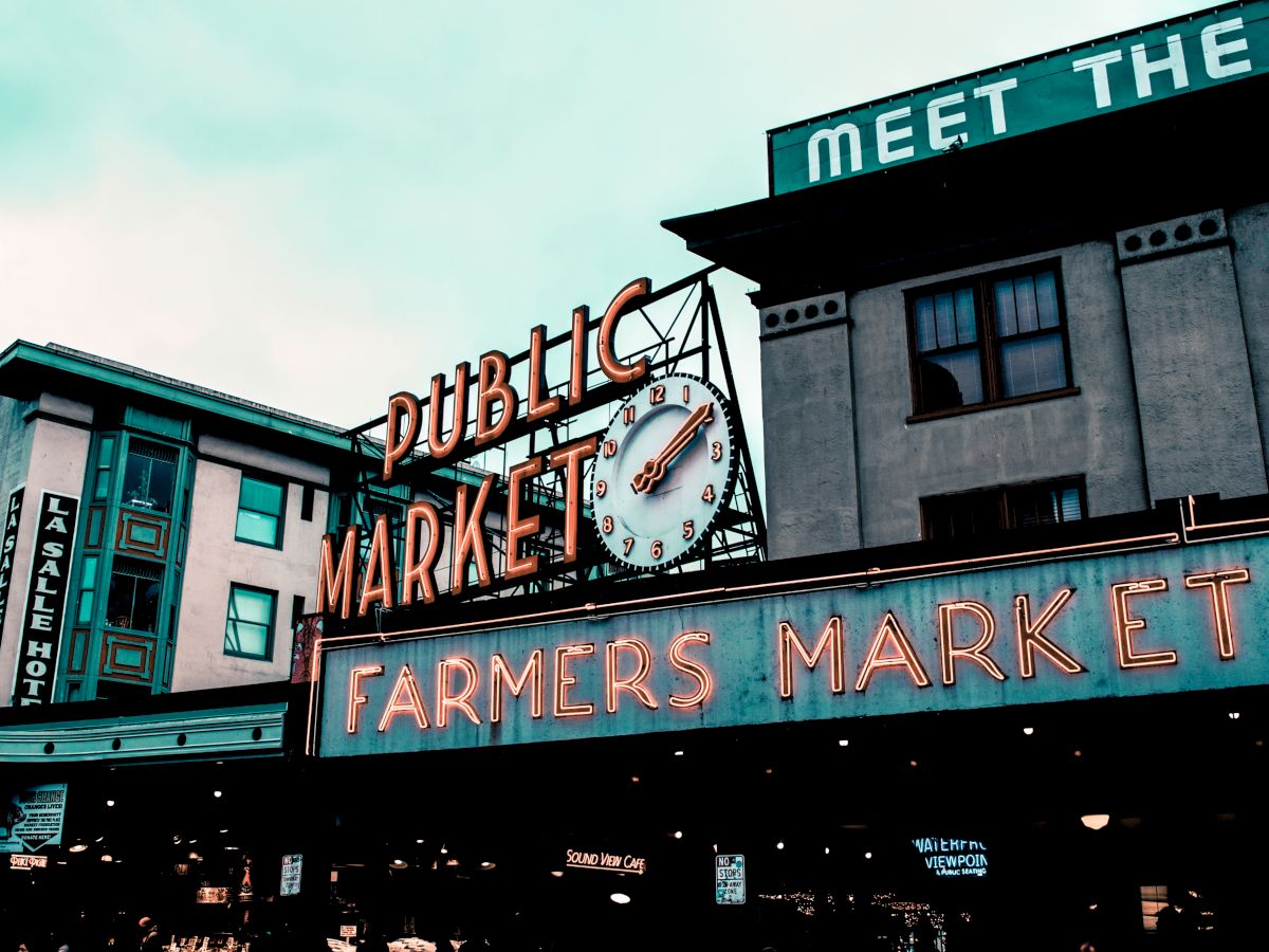 The image shows a public market sign with a clock and a neon "Farmers Market" sign in an urban setting.