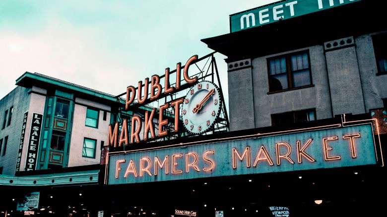 The image shows a public market sign with a clock and a neon "Farmers Market" sign in an urban setting.