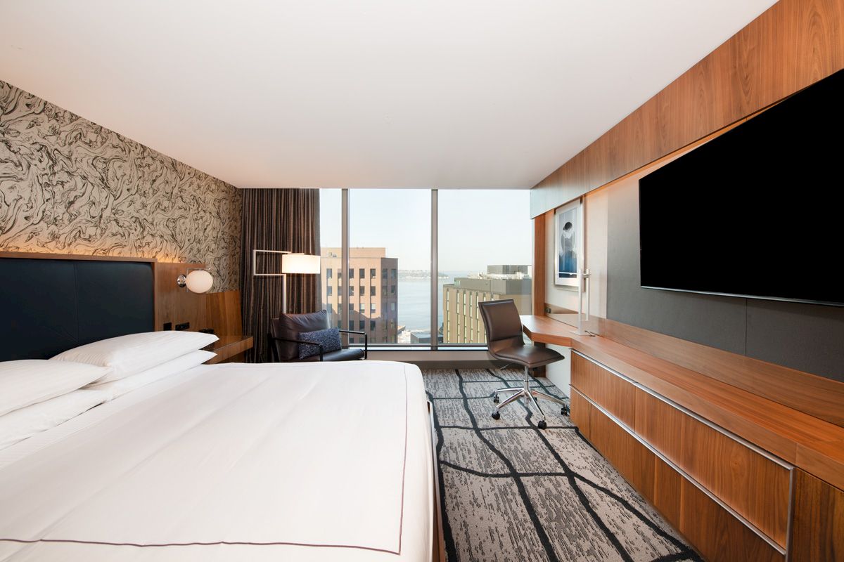 A modern hotel room with a large bed, a desk with a chair, a wall-mounted TV, and windows showing a cityscape view.