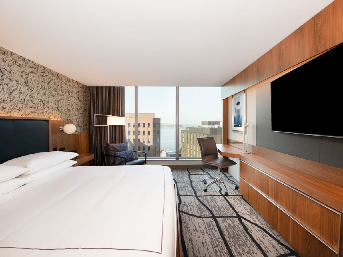 A modern hotel room with a large bed, a desk with a chair, a wall-mounted TV, and windows showing a cityscape view.