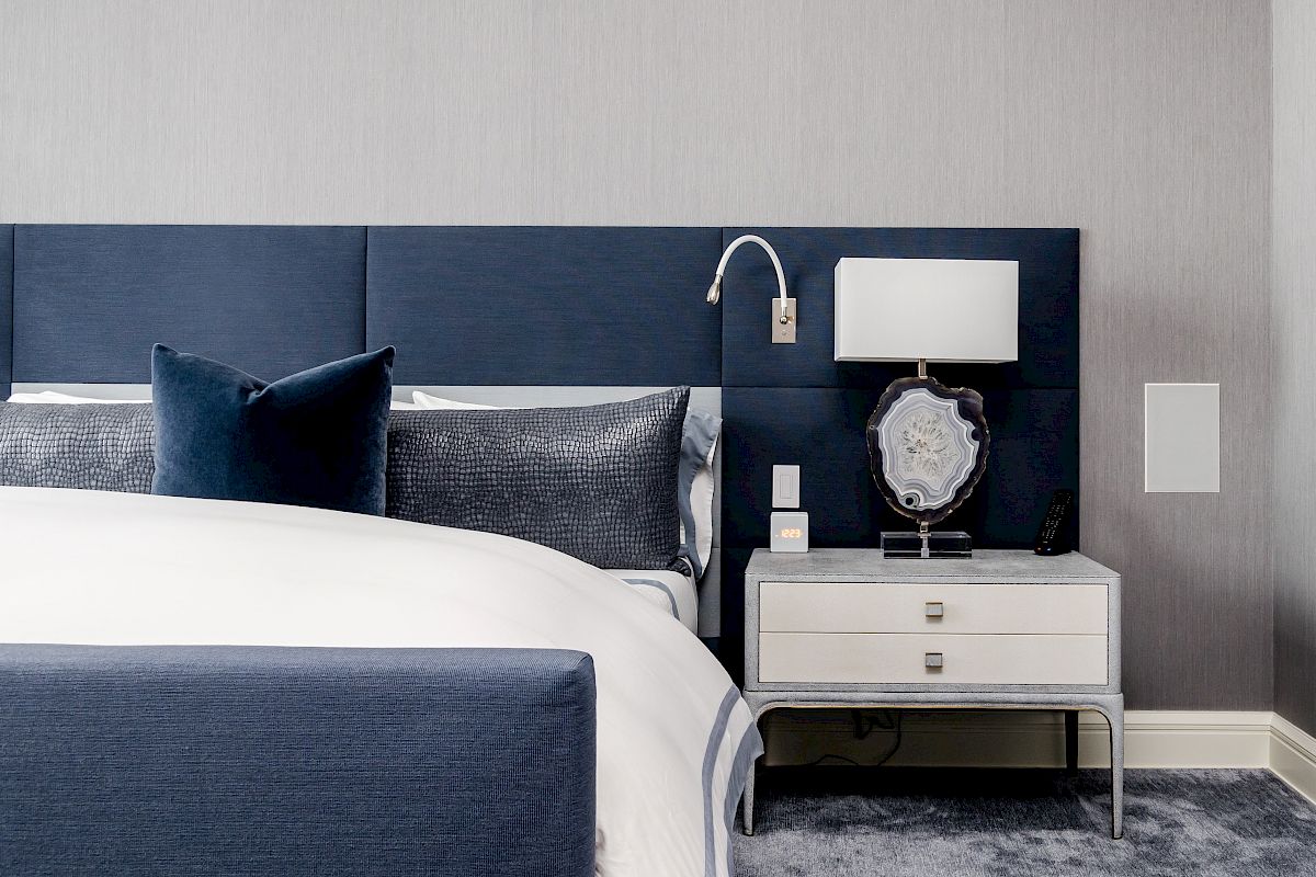 A modern bedroom with a navy blue upholstered bed, dark pillows, a nightstand with a lamp and decor, and a gray carpeted floor.
