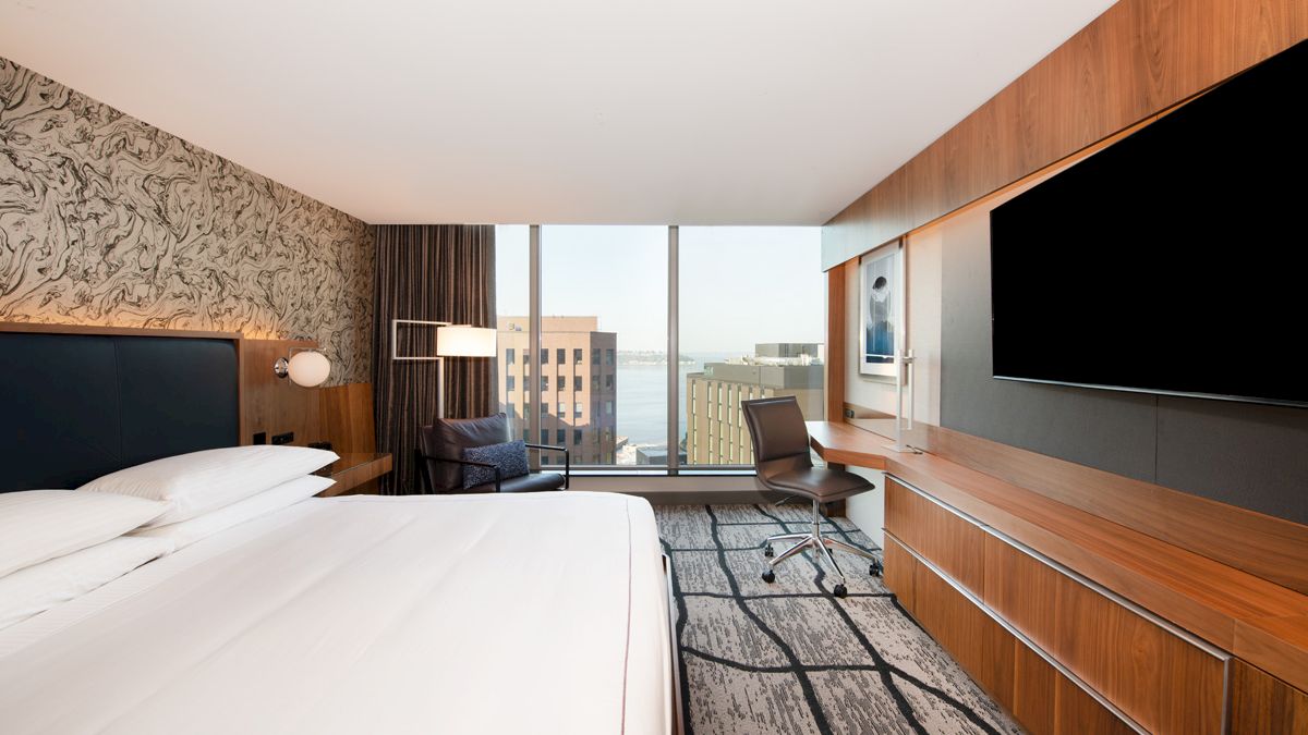 A modern hotel room with a large bed, desk, flat-screen TV, and a window view of city buildings. Brown tones and patterned carpet.