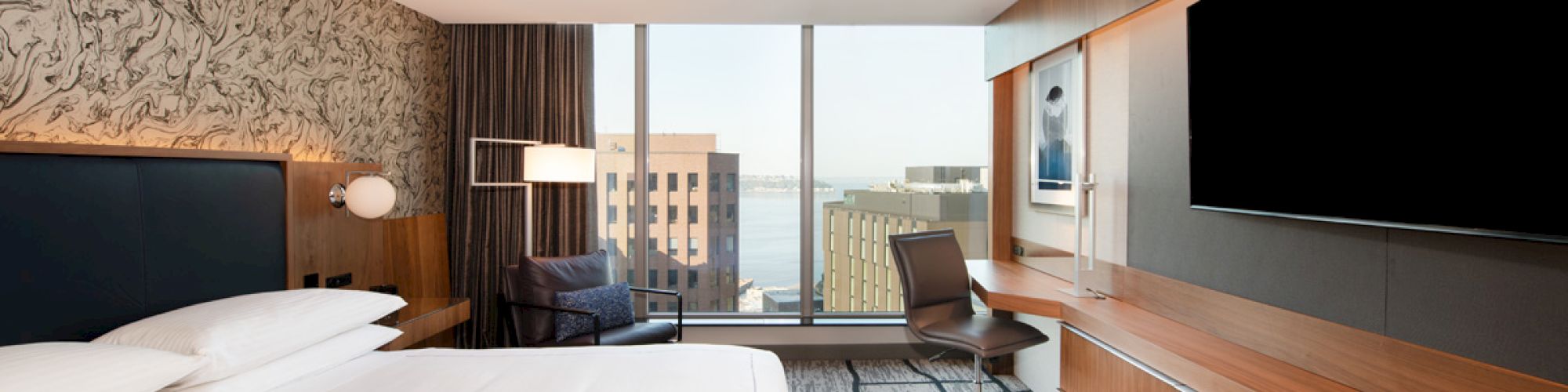 A modern hotel room with a large bed, desk, flat-screen TV, and a window view of city buildings. Brown tones and patterned carpet.